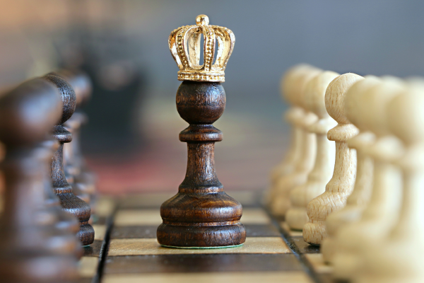 a chessboard with pawns lined up on each side of one in focus centered with gold crown