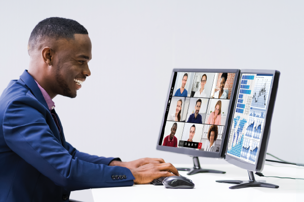 a happy person looking at two computer screens, one with a video conferencing screen showing multiple faces and one showing graphs less visible