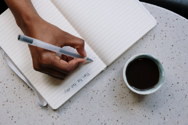 an aerial view of a person's hand writing in a notebook next to a cup of coffee
