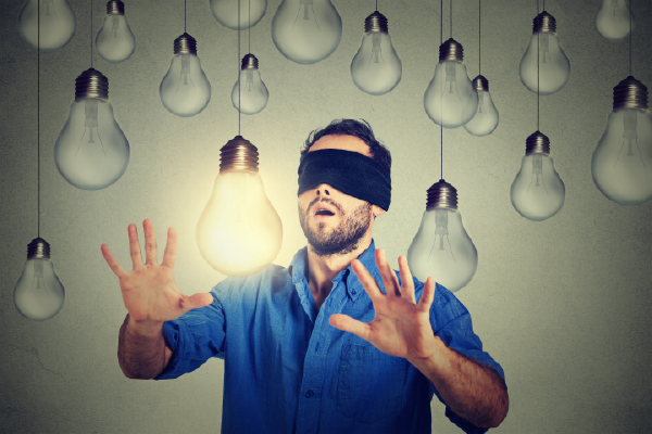 an image of a blindfolded person with their hands in the air near a lit lightbulb surrounded by many dim lightbulbs