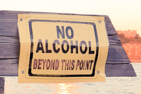 an image of a sign that says 'no alcohol beyond this point' on a wood railing with a sunset reflected in the water below