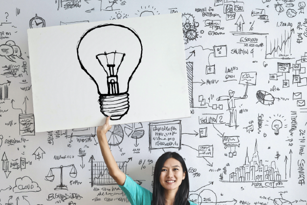 a person in front of a busy whiteboard holding a drawing of a lightbulb above their head