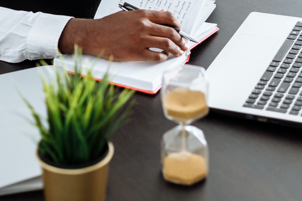 an image of an hour glass on a desk with a person's hand taking notes